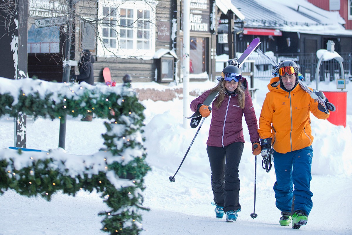 Two skiers walk towards as through a snowy village