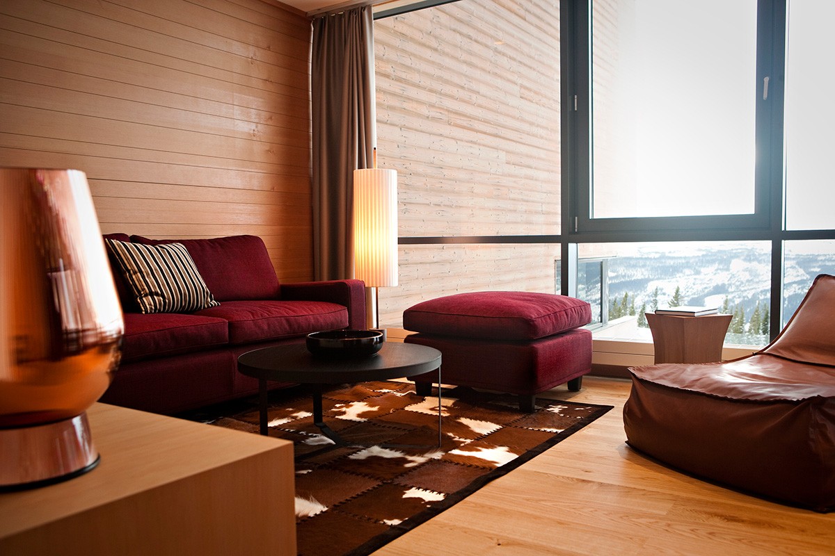 Hotel living area with a red sofa against wood panel walls, beside a large window overlooking a snowy scene